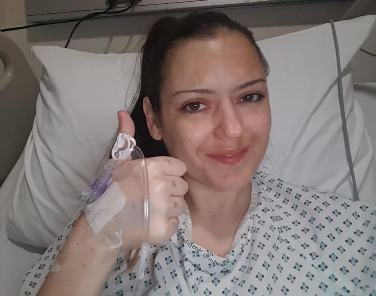 Sahara wearing a hospital gown, in a hospital bed, with canula attached to her hand, giving thumbs up and smiling