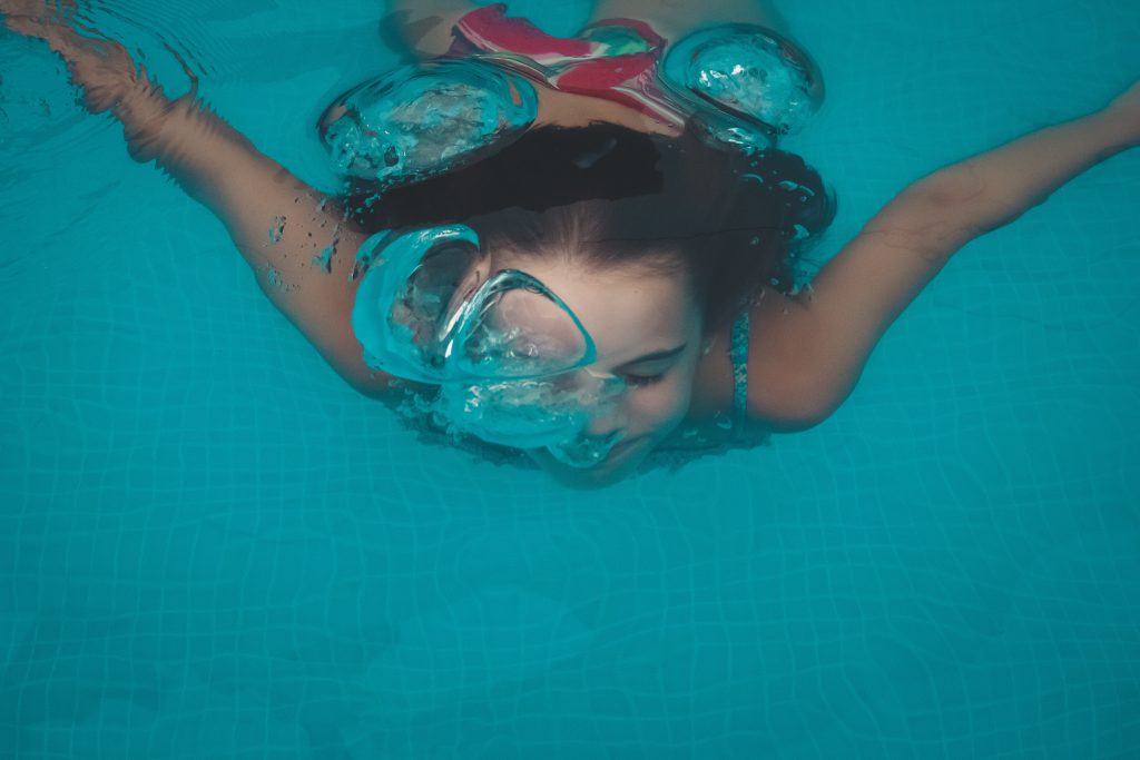 Young woman in a red swimming costume, swimming towards camera under water, blowing bubbles and smiling