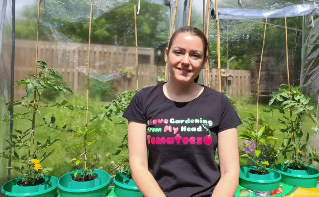 Sahara sits inside a greenhouse with tomato plants, wearing a t-shirt that reads: I love gardening from my head tomatoes