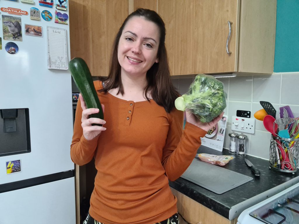 Sahara in her kitchen wearing a russet coloured top, smiling and holding a courgette in one hand and a stem of broccoli in the other.