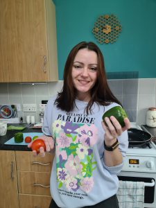 Sahara in her kitchen smiling and holding a red pepper in her right hand and a green pepper in her left hand.