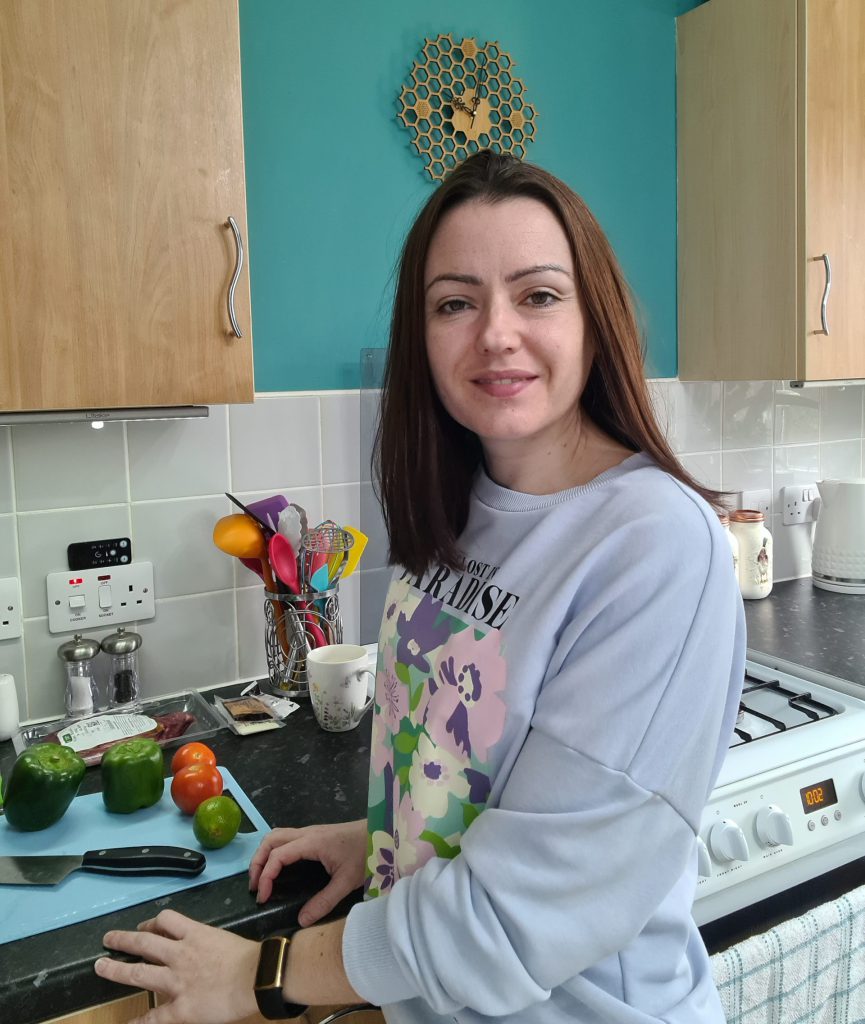 Sahara standing at her kitchen worktop, turning to camera and smiling, with red and green peppers on a chopping board.