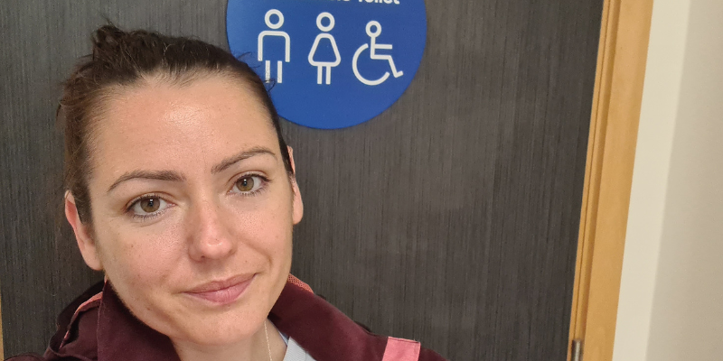 Sahara stands outside a public toilet door with a sign showing icons depicting a man, a woman, and a person in a wheelchair
